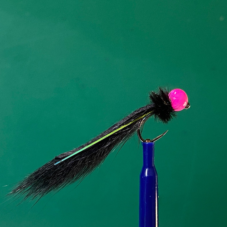 May include: A close-up of a fishing fly with a black body, a pink bead head, and a curved hook. The fly has a black, feathery body with a thin, yellow and blue accent. The hook is held by a blue tool against a green background.