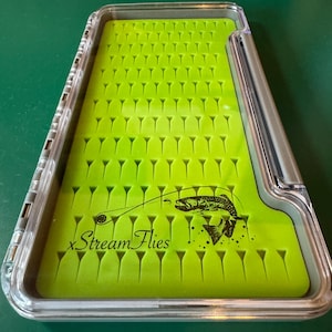May include: A clear plastic fly fishing box with a bright green foam interior. The foam is cut with numerous slots for holding flies. The box has a hinged lid and the words "Stream Flies" are printed on the interior.