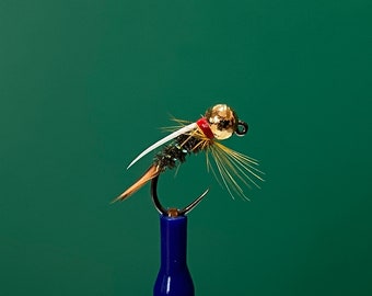May include: A close-up of a fishing fly with a gold-colored bead head, black hook, and various colored feathers. The fly has a red band near the head, white wing-like structures, and brown and yellow accents. The fly is set against a green background.