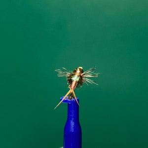 May include: A gold and green fly fishing lure with copper wire legs and a green body. The lure is mounted on a blue fly tying tool.