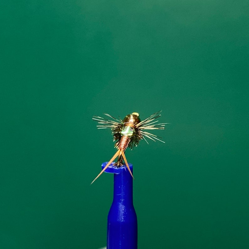 May include: A gold and green fly fishing lure with copper wire legs and a green body. The lure is mounted on a blue fly tying tool.