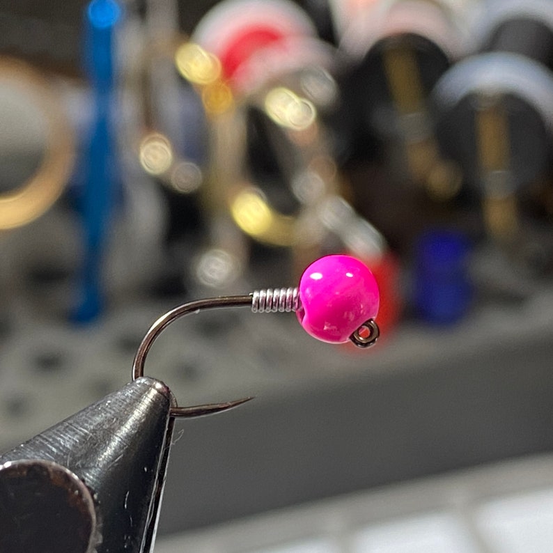 May include: Close-up of a fishing jig with a bright pink, round head and a silver hook. The head is attached to the hook with a silver wire coil. The hook is held in a vise, with other fishing supplies in the blurred background.