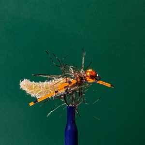 May include: A fishing fly lure featuring a metallic orange bead head, a textured light tan body, and long orange rubber legs with black tips. The fly is constructed with dark, wispy synthetic fibers and is mounted on a sharp metal hook.