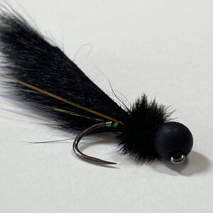May include: A close-up of a black fishing fly with a curved hook. The fly features a black, fluffy body and tail, with accents of gold, green, and purple. The fly is designed for catching fish.