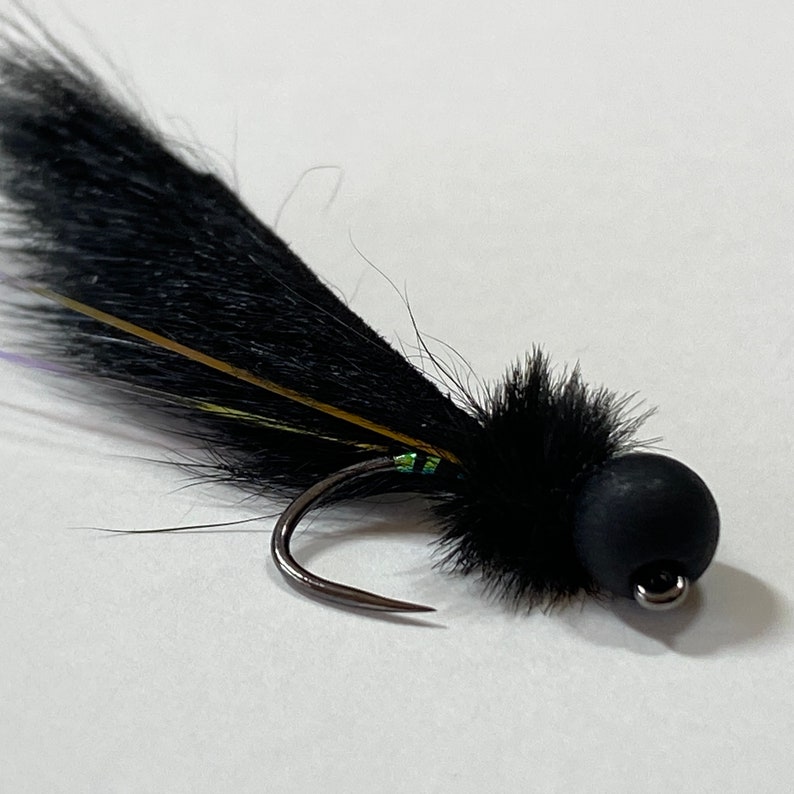 May include: A close-up of a black fishing fly with a curved hook. The fly features a black, fluffy body and tail, with accents of gold, green, and purple. The fly is designed for catching fish.