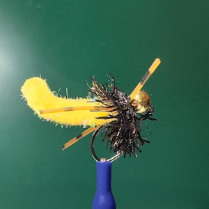 K&ouml;nnte beinhalten: Eine gelb-schwarze Fliegenfischer-K&ouml;der mit einem goldfarbenen Perlenkopf und schwarzen Akzenten. Der K&ouml;der hat einen Haken und ist an einer blauen Basis befestigt. Der K&ouml;der ist so gestaltet, dass er einem Insekt &auml;hnelt.