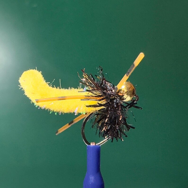 K&ouml;nnte beinhalten: Eine gelb-schwarze Fliegenfischer-K&ouml;der mit einem goldfarbenen Perlenkopf und schwarzen Akzenten. Der K&ouml;der hat einen Haken und ist an einer blauen Basis befestigt. Der K&ouml;der ist so gestaltet, dass er einem Insekt &auml;hnelt.