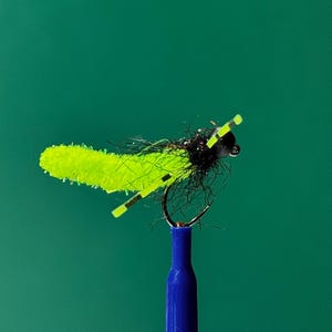 Puede incluir: Un señuelo de pesca con mosca de color verde brillante con fibras negras y un anzuelo. El señuelo está montado en una caña azul.