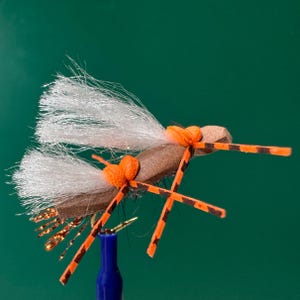 Puede incluir: Primer plano de una mosca de pesca artesanal. Presenta un cuerpo marrón, alas blancas y patas a rayas naranjas y negras. La mosca está montada sobre un soporte azul sobre un fondo verde.