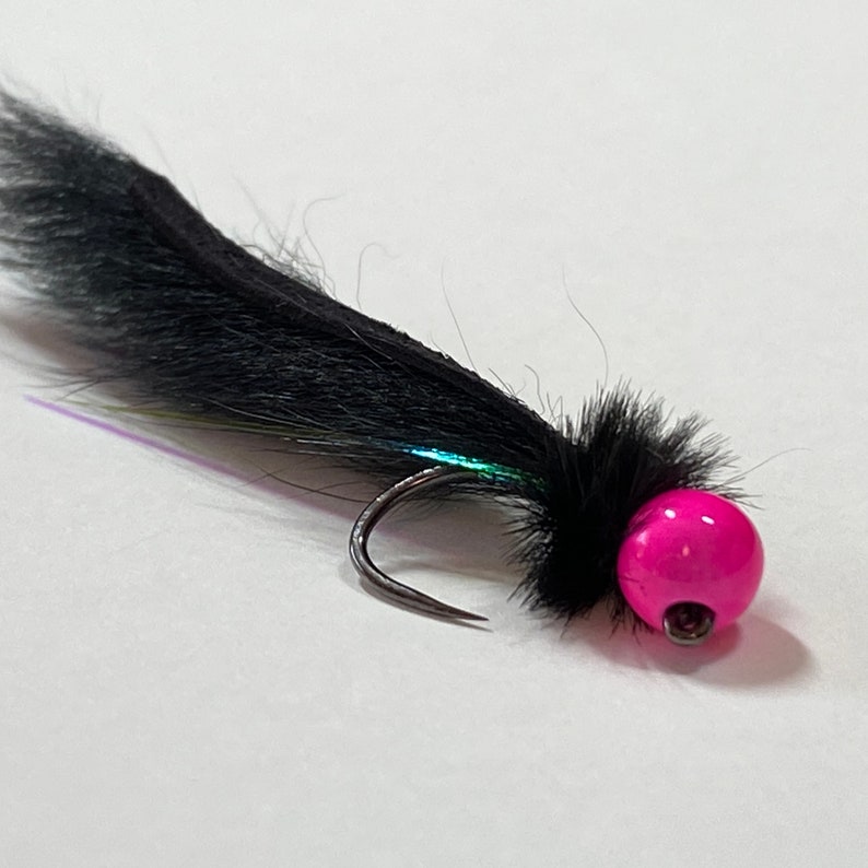 May include: A close-up of a fishing fly with a black, fluffy body and a bright pink bead head. The hook is silver, and the fly has iridescent accents.