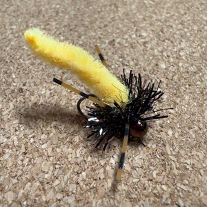 K&ouml;nnte beinhalten: Eine gelb-schwarze Angel-Fliege mit einem flauschigen K&ouml;rper und schwarzen und braunen Akzenten. Die Fliege hat einen Haken und ist so konzipiert, dass sie ein Insekt nachahmt. Die Fliege ist f&uuml;r das Fliegenfischen konzipiert.