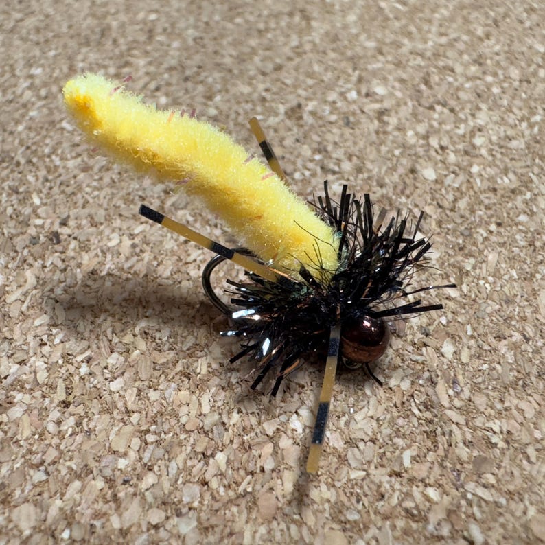 K&ouml;nnte beinhalten: Eine gelb-schwarze Angel-Fliege mit einem flauschigen K&ouml;rper und schwarzen und braunen Akzenten. Die Fliege hat einen Haken und ist so konzipiert, dass sie ein Insekt nachahmt. Die Fliege ist f&uuml;r das Fliegenfischen konzipiert.