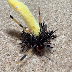K&ouml;nnte beinhalten: Eine gelb-schwarze Angel-Fliege mit einem flauschigen K&ouml;rper und schwarzen Borsten. Die Fliege hat drei beigefarbene Beine mit schwarzen Streifen und eine kleine, runde, kupferfarbene Perle.