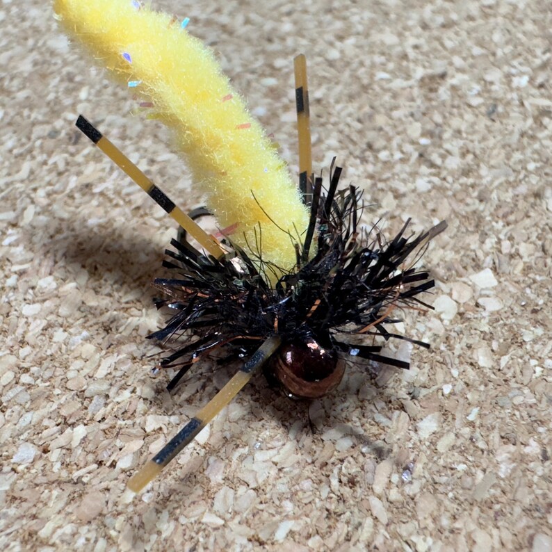 K&ouml;nnte beinhalten: Eine gelb-schwarze Angel-Fliege mit einem flauschigen K&ouml;rper und schwarzen Borsten. Die Fliege hat drei beigefarbene Beine mit schwarzen Streifen und eine kleine, runde, kupferfarbene Perle.