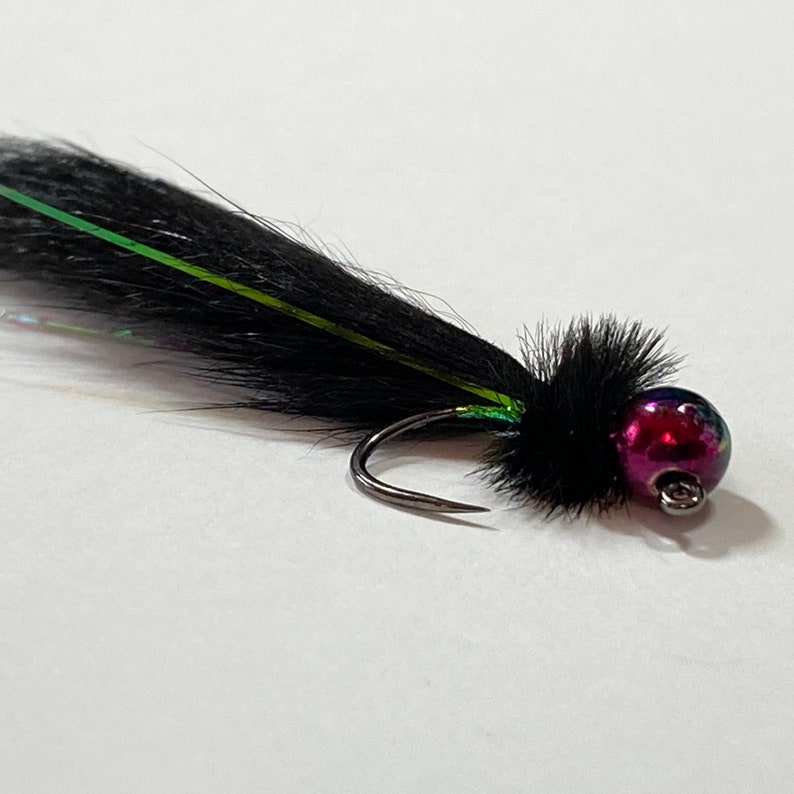 May include: A close-up of a black fishing fly with a curved hook. The fly features a black feathery body with iridescent green accents and a small, round, purple and red bead head. The fly is designed for fishing.