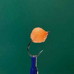 May include: A close-up of a peach-colored fly fishing lure with a black hook and a blue handle.