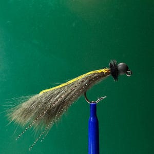 Puede incluir: Una mosca de pesca hecha a mano con cuerpo gris, detalles amarillos y cabeza negra. El anzuelo plateado está fijado a un soporte azul, sobre un fondo verde. Esta mosca está diseñada para la pesca.