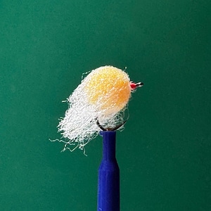 May include: A close-up of a fishing fly with a yellow, fluffy body and white, wispy material. The fly is attached to a black hook and a blue handle, set against a green background.