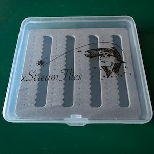 Könnte beinhalten: Eine transparente Fliegenfischerbox mit grauem Schaumstoff-Innenraum. Die Box hat einen Klappdeckel und zeigt den Text "x Stream Flies" sowie eine Grafik eines Fisches mit einer Angelschnur.