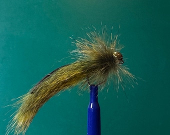 Può includere: Un'esca artificiale da pesca con un corpo lungo e affusolato realizzato in fibre sintetiche color verde oliva e marrone. La testa dell'esca è composta da fibre marroni dense e spinose con un piccolo occhio a perla metallica riflettente.