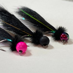 May include: Close-up of three fishing flies. Each fly features a black, feathery body and a colorful, iridescent bead head. One fly has a bright pink bead, another a black bead, and the third a multi-colored bead. The hooks are silver.