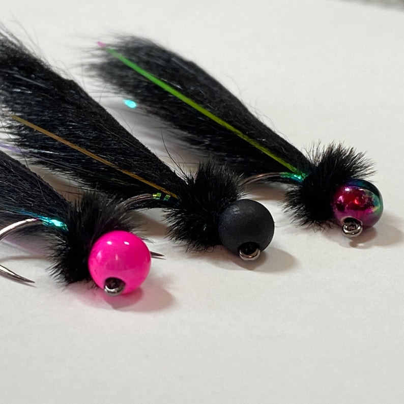 May include: Close-up of three fishing flies. Each fly features a black, feathery body and a colorful, iridescent bead head. One fly has a bright pink bead, another a black bead, and the third a multi-colored bead. The hooks are silver.