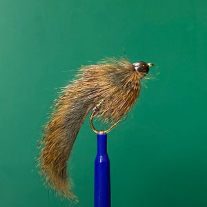 May include: A brown and gray fly fishing lure with a dark brown head and a gold hook. The lure is mounted on a blue rod.