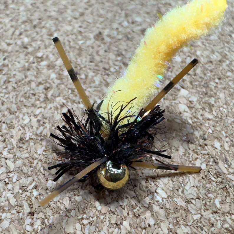 K&ouml;nnte beinhalten: Nahaufnahme einer gelb-schwarzen Fliegenfischer-K&ouml;der. Der K&ouml;der hat einen gelben, flauschigen K&ouml;rper, schwarze und braune Akzente und eine goldfarbene Perle. Der K&ouml;der hat mehrere braune und schwarze Anh&auml;nge, die sich nach au&szlig;en erstrecken.