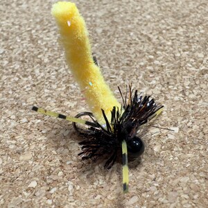 K&ouml;nnte beinhalten: Eine gelb-schwarze Angel-Fliege mit einem flauschigen gelben K&ouml;rper und schwarzen Akzenten. Die Fliege hat einen schwarzen Kopf und zwei gelb-schwarz gestreifte Anh&auml;nge. Die Fliege ist zum Fangen von Fischen bestimmt.
