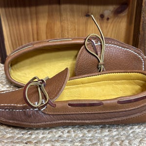 May include: Brown leather moccasin-style shoes with white stitching and yellow insoles. The shoes have a classic design with a leather lace detail. The second shoe is in the background, showing the inside of the shoe.