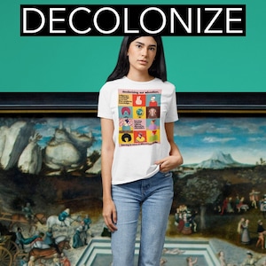 May include: A white t-shirt with a graphic print of a colorful illustration that says "Decolonizing our education." The illustration features diverse people and symbols representing different cultures and knowledge systems. The shirt is worn by a person standing in front of a painting.