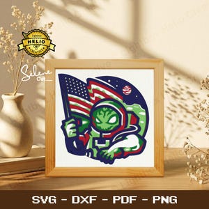 May include: A framed graphic of an alien astronaut holding an American flag. The alien is green with a red and white spacesuit. The background is a blue night sky with a planet. The frame is made of light brown wood. The image includes the text "SVG - DXF - PDF - PNG".