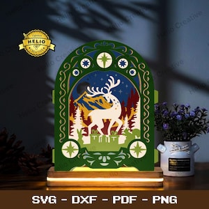 Peut inclure: Une boîte lumineuse arquée à plusieurs couches représentant la silhouette d'un cerf blanc parmi des cadeaux dans une forêt avec des montagnes et un ciel étoilé. Le motif est encadré de vert foncé avec des détails dorés sur un socle en bois.