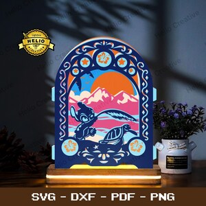Può includere: Una scatola luminosa ad arco multistrato con una cornice blu scuro caratterizzata da ritagli di fiori di ibisco e stelle. Il centro mostra una scena a strati con un personaggio alieno blu e una tartaruga marina su uno sfondo di montagne rosa e sole arancione.
