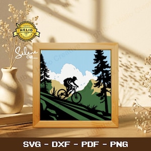 Op de afbeelding: Een ingelijste print met een zwart silhouet van een fietser op een mountainbike, tegen een achtergrond van groene heuvels, bomen en een blauwe lucht met wolken. De lijst is van licht hout. De tekst 'SVG - DXF - PDF - PNG' staat onderaan het frame.