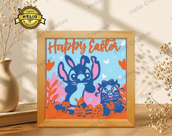 Puede incluir: Un marco de madera cuadrado que contiene una impresión gráfica con el texto Happy Easter. El diseño presenta un personaje azul con orejas largas, una cesta de Pascua, mariposas y huevos estampados sobre un fondo azul claro y rosa.