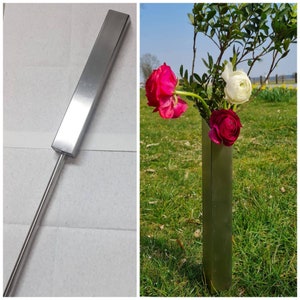 Könnte beinhalten: Eine silberne Metallvase mit quadratischer Form, die in einem grasbewachsenen Feld steht. Die Vase enthält drei Blumen: eine rosa Blume, eine weiße Blume und eine rote Blume.