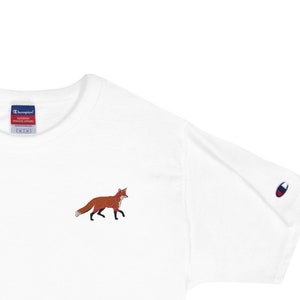 Könnte beinhalten: Weißes T-Shirt mit kurzen Ärmeln und einem kleinen gestickten Fuchs-Design auf der linken Brust. Das Shirt hat ein Champion-Markenetikett am linken Ärmel.