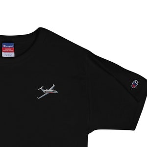 May include: Black t-shirt with a small embroidered image of a private jet on the chest. The shirt has a crew neck and short sleeves. The Champion logo is visible on the left sleeve.