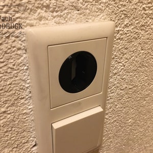 Könnte beinhalten: Eine cremefarbene Steckdose und ein Schalter sind an einer strukturierten Wand montiert. Die Steckdose hat eine große, runde, schwarze Öffnung. Der Schalter darunter ist rechteckig und geschlossen. Der Text "Fach-AusDRUCK" befindet sich in der oberen linken Ecke.