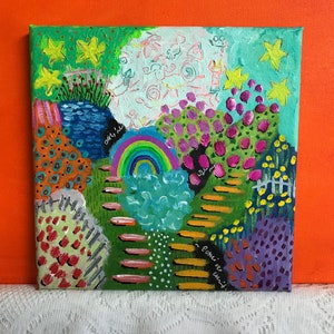 May include: Square acrylic painting depicting a whimsical garden scene. The artwork features a rainbow, stepping stones, and various colorful plants and flowers. Yellow stars and a white picket fence add to the playful atmosphere. The painting is set against an orange background.
