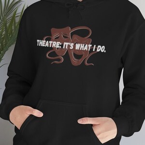 May include: Black hoodie with the text "THEATRE: IT'S WHAT I DO." and a graphic of two brown theatre masks. The hoodie has a front pocket and a drawstring hood. A casual garment for theatre enthusiasts.