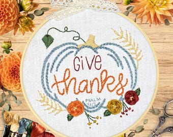 May include: Embroidered hoop art featuring a pumpkin design and the words "Give Thanks." The pumpkin is outlined in blue with orange lettering. Floral accents in orange, yellow, and red surround the pumpkin. A white lace bow is at the top.