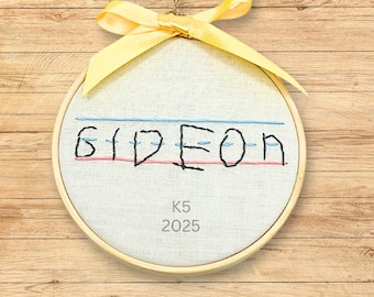 May include: A circular wooden embroidery hoop containing off-white fabric with the name GIDEON embroidered in black thread between blue and pink lines. Below the name, the text K5 2025 is printed. A yellow satin ribbon is tied at the top.