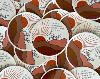 Può includere: Una collezione di adesivi rotondi con un design a forma di alba. Gli adesivi presentano un sole rosso, montagne marroni e la scritta "In the morning when I rise give me Jesus" in stile scritto a mano.