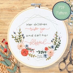 Może przedstawiać: Haftowany obręcz z kwiatowym wiankiem i cytatem "Jej dzieci wstają i nazywają ją błogosławioną" z odniesieniem do Pisma Świętego "Prz 31:28".