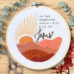 May include: Embroidered hoop art with a sunrise over mountains and the text "In the morning when I rise give me Jesus".