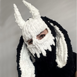 Puede incluir: Una máscara de punto blanca y negra con largas y esponjosas orejas blancas. La máscara tiene un diseño de calavera y cubre la cara.