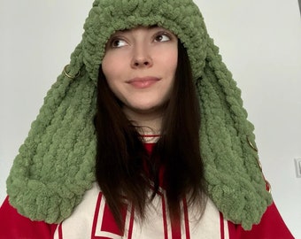 May include: A sage green chunky knit hat with long, dangling ear-like flaps. The hat features a thick, textured yarn construction and is accented with small, circular gold-toned metal rings attached to the sides of the long, hanging panels.