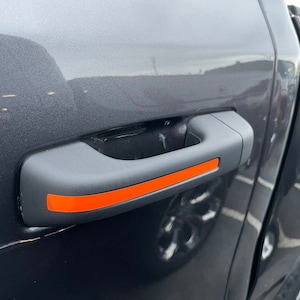 May include: A black car door handle with an orange stripe. The handle is made of plastic and has a smooth surface.